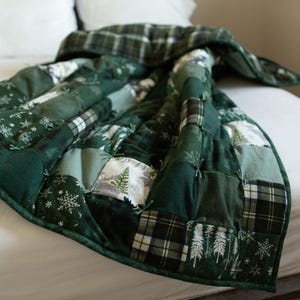 May include: A patchwork quilt featuring various shades of green, including forest and sage. The squares display patterns of plaid, snowflakes, and pine trees. The quilt is constructed with visible tied seams throughout the fabric surface.