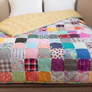 May include: A colorful patchwork quilt with a variety of fabrics and patterns. The quilt is made up of squares of different colors and designs, including floral, geometric, and plaid patterns. The quilt is laid out on a bed with a yellow bedspread underneath.