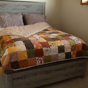 May include: A patchwork quilt featuring a grid of square fabric patches in various shades of brown, orange, yellow, and green with floral and geometric patterns. The quilt is draped over a bed with a light tan underside and a matching border.