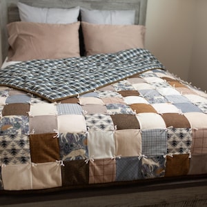 Puede incluir: Un edredón de retazos con una variedad de patrones cuadrados en tonos marrones, beige y azul. El edredón presenta diseños de ciervos y geométricos. Una manta a cuadros y dos almohadas rosas están en la cama.