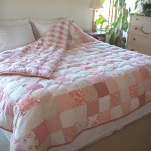 May include: A pink and white patchwork quilt with a ruffled edge. The quilt is made of various fabrics with different patterns and textures. It is laid out on a bed with white sheets and pillows.