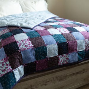 May include: A rag quilt featuring a patchwork design of square fabric pieces in shades of purple, navy blue, teal, and white. The squares are joined with exposed seams and tied with small fabric knots, creating a textured surface across the bedding.