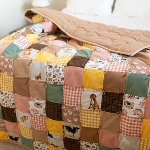 May include: A patchwork quilt with a variety of fabrics in shades of yellow, brown, pink, and green. The quilt features squares with dog and farm animal patterns. The top layer is a quilted, solid tan fabric.