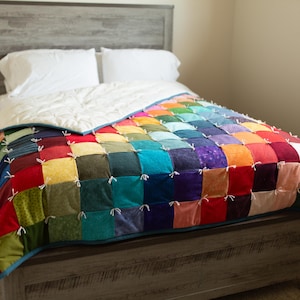 May include: A bed with a vibrant patchwork quilt. The quilt is made up of squares in a variety of colours, such as red, orange, yellow, green, blue, and purple. White pillows and a white comforter are on the bed. The bed frame is wooden.