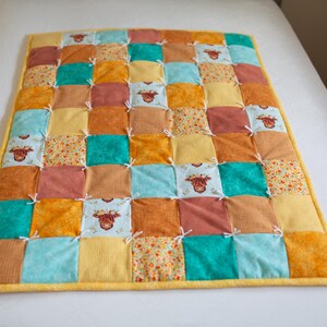 May include: A patchwork quilt with a yellow border. The quilt is made of squares in shades of orange, yellow, teal, and brown. Some squares feature a cow illustration. White ties secure the squares.