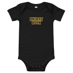 Puede incluir: Body de bebé negro con el texto "PHIRST BORN" en una fuente dorada y negra de estilo universitario. El body tiene mangas cortas y corchetes en la parte inferior.