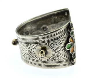 May include: Silver-toned cuff bracelet with intricate floral and geometric patterns. The bracelet features a decorative flower with green, yellow, and orange accents. The cuff has a curved design and a small, round knob on the side.