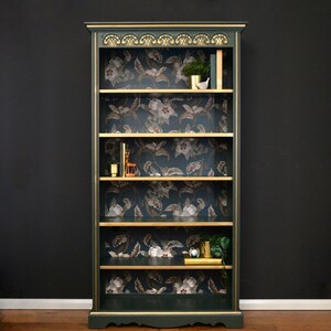 Puede incluir: Una librer&iacute;a verde oscuro con detalles dorados y cinco estantes. Los estantes est&aacute;n forrados con papel tapiz floral. La librer&iacute;a tiene una base festoneada y una parte superior decorativa.