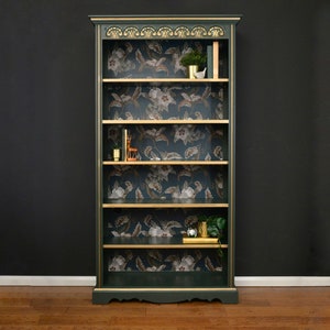 Puede incluir: Una librer&iacute;a verde oscuro con molduras doradas y cinco estantes. Los estantes est&aacute;n forrados con papel tapiz floral y decorados con peque&ntilde;as plantas y objetos decorativos.