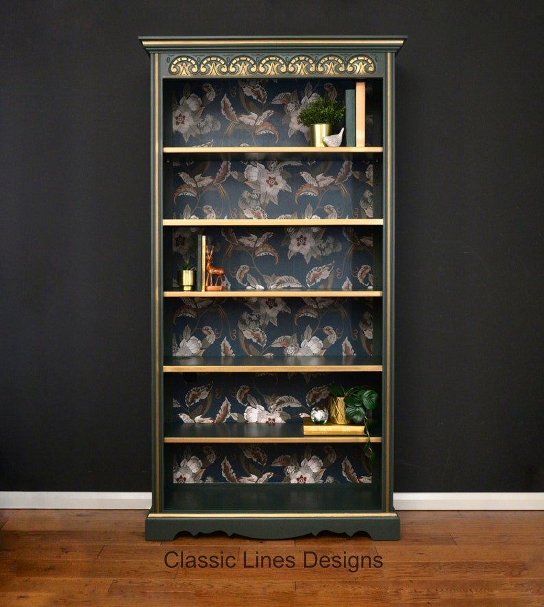 Puede incluir: Una librer&iacute;a verde oscuro con detalles dorados y cinco estantes. Los estantes est&aacute;n forrados con papel tapiz floral. La librer&iacute;a tiene una base festoneada y una parte superior decorativa.