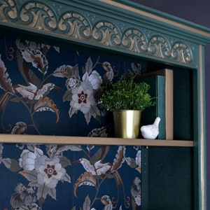 Puede incluir: Una estanter&iacute;a de madera pintada de verde con molduras doradas y un papel tapiz floral que recubre los estantes. Una peque&ntilde;a planta en maceta y una figurita de p&aacute;jaro de cer&aacute;mica blanca est&aacute;n en el estante superior.