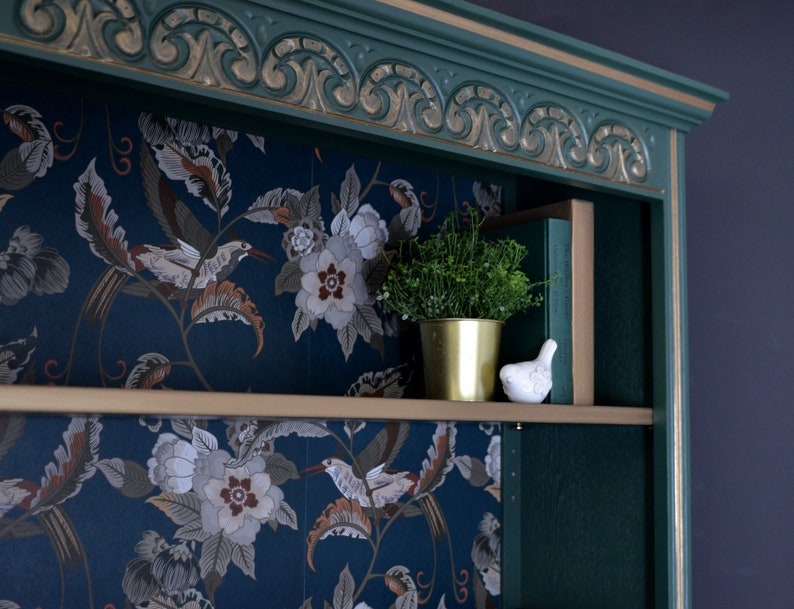 Puede incluir: Una estanter&iacute;a de madera pintada de verde con molduras doradas y un papel tapiz floral que recubre los estantes. Una peque&ntilde;a planta en maceta y una figurita de p&aacute;jaro de cer&aacute;mica blanca est&aacute;n en el estante superior.