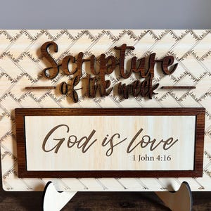 May include: Wooden sign with the text "Scripture of the week" in a dark brown, raised font. Below, a rectangular frame contains the words "God is love" and the Bible verse 1 John 4:16.