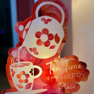 May include: A decorative night light featuring a floral design with a teapot pouring into a teacup. The night light has the text "Tea Time is my HAPPY Place" in white script. The background is a watercolor floral pattern in shades of red and orange.