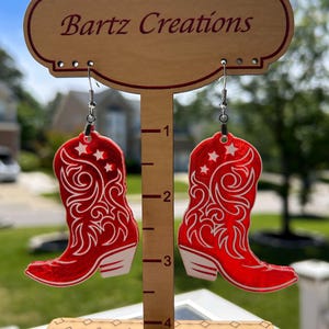 May include: Red and white cowboy boot earrings with a star design. The earrings are hanging from a wooden display stand with the text "Bartz Creations".