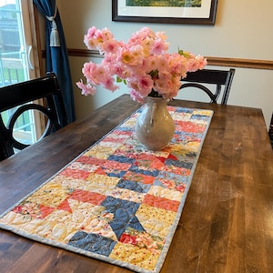 May include: A rectangular quilted table runner featuring a patchwork design of floral and geometric patterns in shades of blue, coral, yellow, and white, with a light blue border, topped with a ceramic vase filled with pink cherry blossoms.