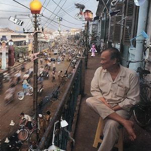 May include: A man sits on a yellow stool on a balcony overlooking a busy street. He is wearing a light brown shirt and pants. The balcony has a metal railing and is decorated with green plants. The street is filled with people and vehicles.