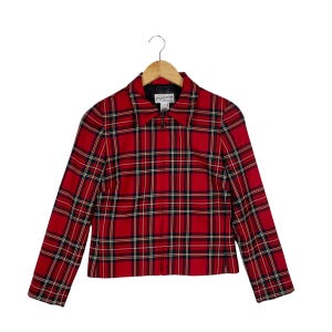 May include: A red, black, and white tartan jacket featuring a full-length front zip, a pointed collar, and long sleeves. The internal label reads Pendleton. The garment is displayed on a wooden hanger against a plain white background.