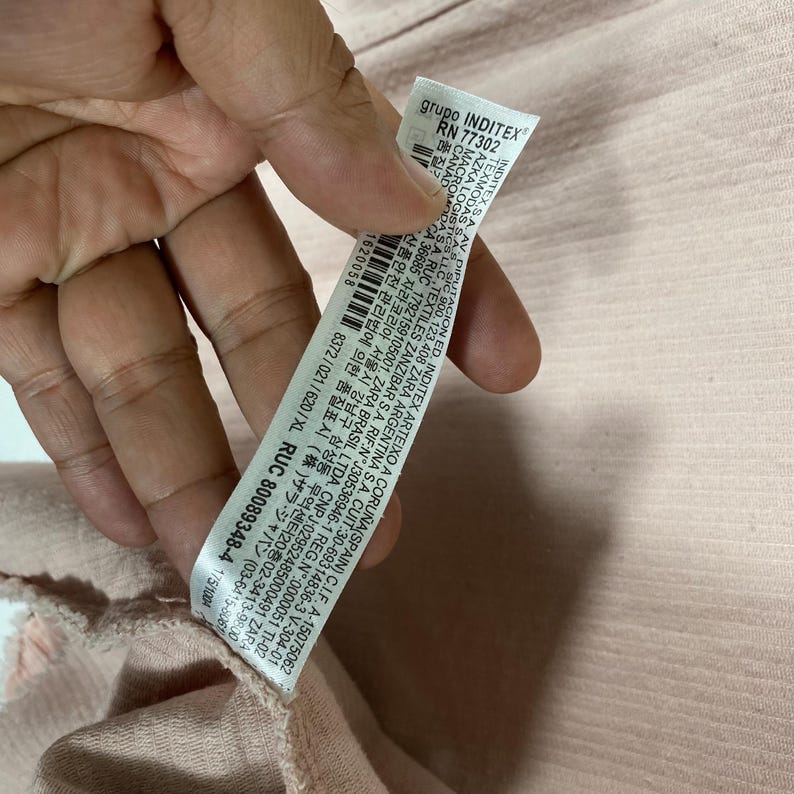 K&ouml;nnte beinhalten: Nahaufnahme eines Kleidungsst&uuml;cketiketts, das von einer Hand gehalten wird. Das Etikett ist wei&szlig; mit schwarzem Text, einschlie&szlig;lich des Markennamens "grupo INDITEX" und der RN-Nummer 77302. Das Etikett enth&auml;lt auch Gr&ouml;&szlig;enangaben, "XL". Der Stoff ist hellrosa.