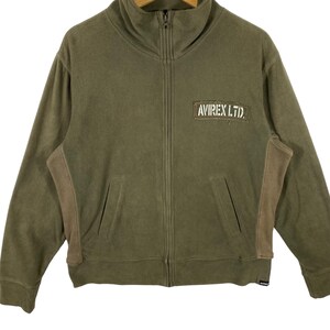 Puede incluir: Chaqueta verde oliva con cremallera, cuello alto y dos bolsillos delanteros. La chaqueta presenta paneles marrones en los laterales y un parche rectangular con el texto "AVIREX LTD." en blanco. Cremallera completa y etiqueta peque&ntilde;a en la parte inferior.