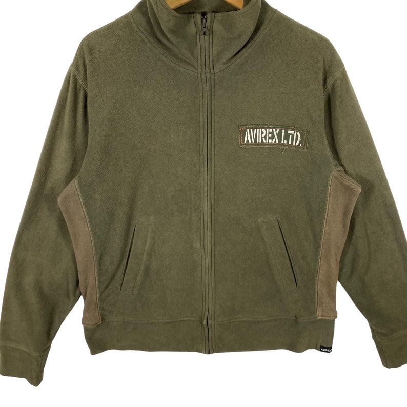 Puede incluir: Chaqueta verde oliva con cremallera, cuello alto y dos bolsillos delanteros. La chaqueta presenta paneles marrones en los laterales y un parche rectangular con el texto "AVIREX LTD." en blanco. Cremallera completa y etiqueta peque&ntilde;a en la parte inferior.
