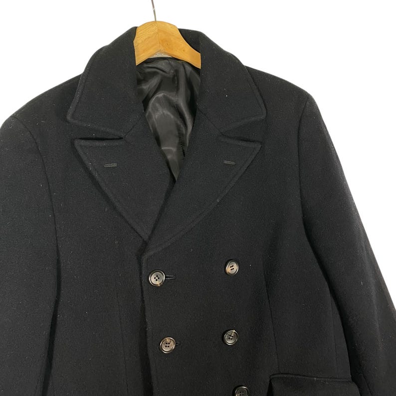 Pu&ograve; includere: Un cappotto nero doppiopetto con revers a lancia e quattro bottoni scuri. Il cappotto &egrave; appeso a una gruccia di legno. Il cappotto ha un design classico e su misura.