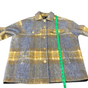 May include: A gray and yellow plaid jacket with gold buttons and two chest pockets. The jacket has a collared neckline and long sleeves. The jacket is measured with a green measuring tape.