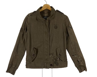 May include: Olive green jacket with a collared neckline, button closures, and multiple pockets. Features patches and printed text details. The jacket is hanging on a wooden hanger.