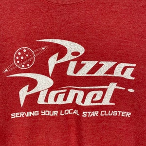 Puede incluir: Camiseta roja con un gr&aacute;fico blanco del logotipo de Pizza Planet, que presenta un planeta con anillos y el texto "Pizza Planet" en una fuente estilizada. El texto "Serving your local star cluster" est&aacute; impreso debajo del logotipo.