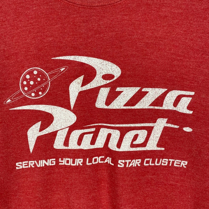 Puede incluir: Camiseta roja con un gr&aacute;fico blanco del logotipo de Pizza Planet, que presenta un planeta con anillos y el texto "Pizza Planet" en una fuente estilizada. El texto "Serving your local star cluster" est&aacute; impreso debajo del logotipo.