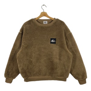 May include: Brown fleece sweatshirt with a crew neck and long sleeves. The sweatshirt features a small black square patch with the text "CAMP7" and a mountain graphic. It is displayed on a wooden hanger.