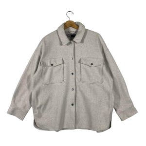 May include: A light gray button-down shirt jacket with two front pockets and black buttons. The jacket is hanging on a wooden hanger. The brand name "ZARA" is visible on the collar.