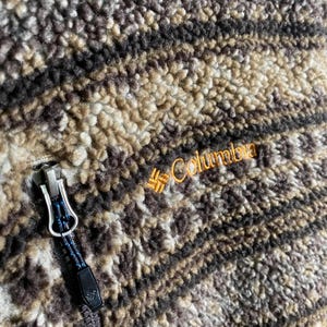 Puede incluir: Una chaqueta de forro polar a rayas marrones y negras con cremallera y el logotipo de Columbia bordado en la parte delantera.