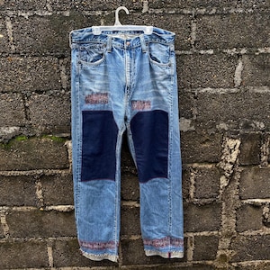 May include: Blue denim jeans with two large dark blue patches on the front. The jeans have a faded look and are distressed.