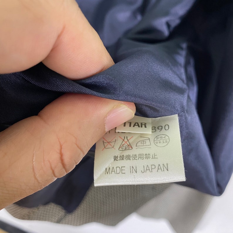 Puede incluir: Una etiqueta de tela azul marino con texto blanco que dice "ITAR 390" y "MADE IN JAPAN". La etiqueta tambi&eacute;n incluye un s&iacute;mbolo de instrucciones de lavado y un s&iacute;mbolo que indica que la tela no debe limpiarse en seco.