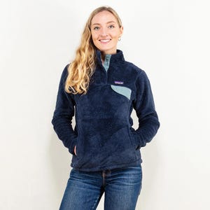 May include: A navy blue fleece pullover with a light blue chest pocket and collar. The pullover has a high collar and a snap closure. The model is wearing blue jeans.