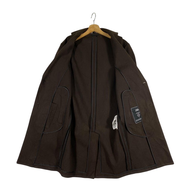 Pu&ograve; includere: Un cappotto lungo con maniche lunghe di colore marrone con chiusura a zip e due tasche interne. Il cappotto ha un'etichetta con la scritta "ZARA" all'interno.