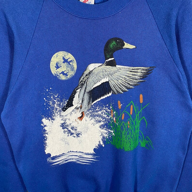K&ouml;nnte beinhalten: Ein blaues Sweatshirt mit einem grafischen Aufdruck eines fliegenden Stockenten-M&auml;nnchens &uuml;ber einem See mit einem Vollmond im Hintergrund. Die Ente ist detailliert dargestellt mit einem gr&uuml;nen Kopf und einer wei&szlig;en Brust.