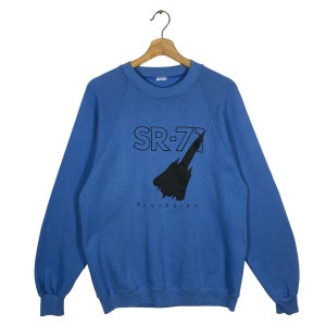 Puede incluir: Una sudadera azul con un gráfico negro de un avión SR-71 Blackbird. El texto "SR-71 BLACKBIRD" está impreso debajo del avión.
