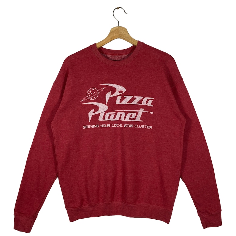 Puede incluir: Sudadera roja con un gr&aacute;fico blanco del logotipo de Pizza Planet de la pel&iacute;cula Toy Story. El logotipo dice "Pizza Planet" y "Serving Your Local Star Cluster".