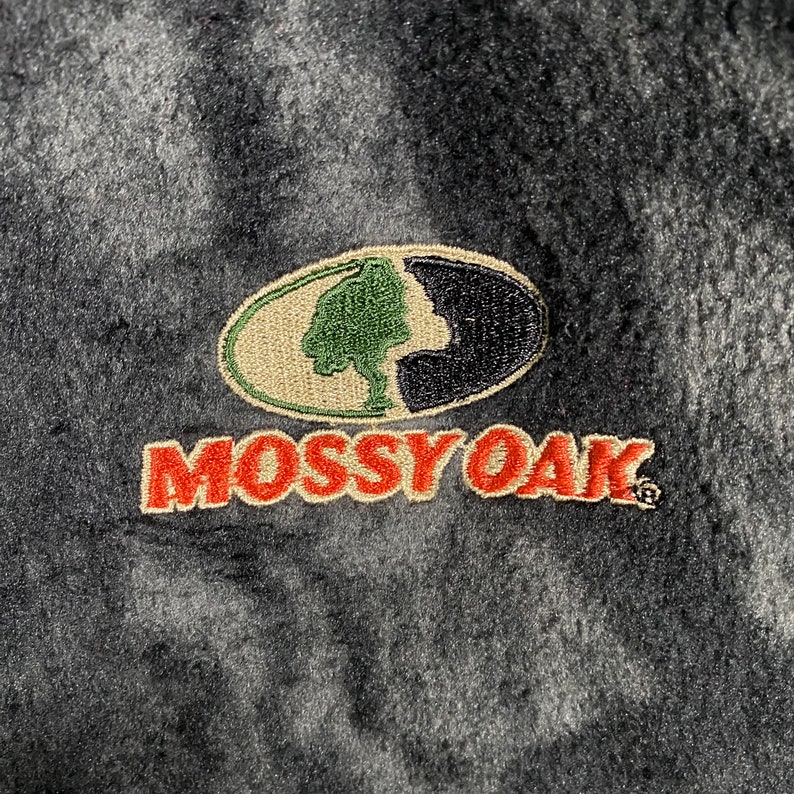 May include: A black and green embroidered patch with a tree and a silhouette of a person's head. The patch is surrounded by a tan oval and the words "Mossy Oak" are embroidered in red below the patch.
