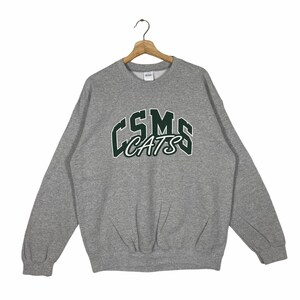 Könnte beinhalten: Grauer Sweatshirt mit einem grün-weißen Logo, auf dem "CSMS CATS" im Retro-Stil steht.