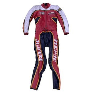 Puede incluir: Un traje de carreras de moto de cuero rojo, blanco y negro con la palabra "Dainese" impresa en las piernas. El traje tiene una cremallera en la parte delantera y un parche blanco en el pecho que dice "Champion".