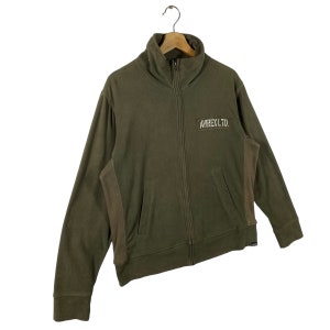 Puede incluir: Chaqueta con cremallera verde oliva con cuello alto y mangas largas. La chaqueta tiene un peque&ntilde;o parche rectangular con el texto "AVIREX L.T.D." en el pecho. Colgada de una percha de madera.