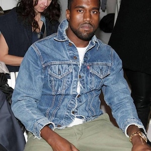 May include: A light blue denim jacket with a button-up front and chest pockets. The jacket is worn over a white t-shirt and paired with olive green pants. A gold watch is visible on the wrist.