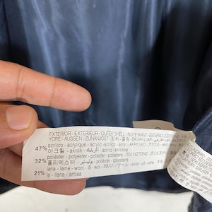 May include: A close-up of a clothing tag with text in multiple languages. The tag lists the exterior shell composition: 47% acrylic, 32% polyester, and 21% lana. The tag is white with black text and is attached to a navy blue garment.