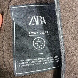 Pu&ograve; includere: Un'etichetta in bianco e nero con il testo "ZARA 4 WAY COAT" e una descrizione delle caratteristiche del cappotto. L'etichetta &egrave; attaccata a un tessuto marrone.
