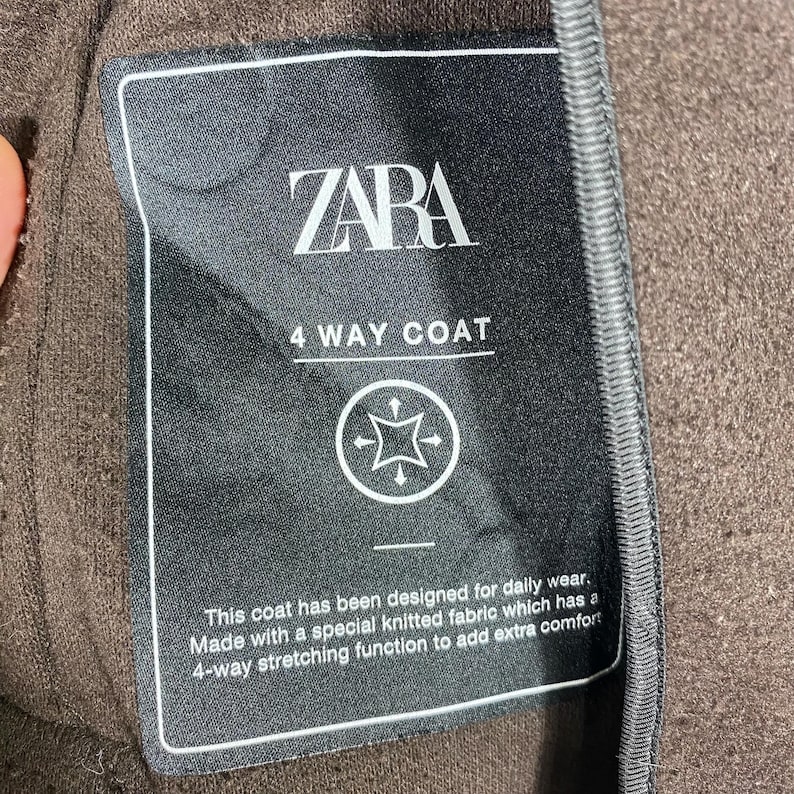Pu&ograve; includere: Un'etichetta in bianco e nero con il testo "ZARA 4 WAY COAT" e una descrizione delle caratteristiche del cappotto. L'etichetta &egrave; attaccata a un tessuto marrone.