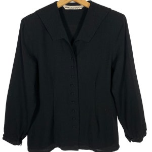 Puede incluir: Una camisa negra de manga larga con botones y cuello. La camisa tiene una silueta ajustada y una etiqueta que dice "COMME des GARCONS".