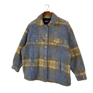 May include: A blue and yellow plaid jacket with a collared neckline, button-down front, and two chest pockets. The jacket is hanging on a wooden hanger. The brand label "ZARA" is visible on the collar.